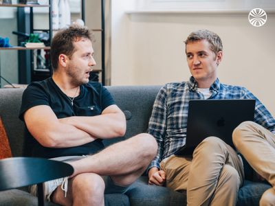 Two white men sitting on a couch in discussion, one holding a laptop.