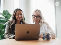 Two women, one with glasses, work together on a laptop at a table, with a multiverse-branded mug nearby.