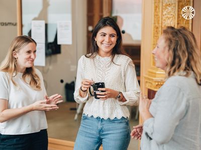 Three white women engaged in a friendly conversation, smiling, holding coffee cups in an office hallway.