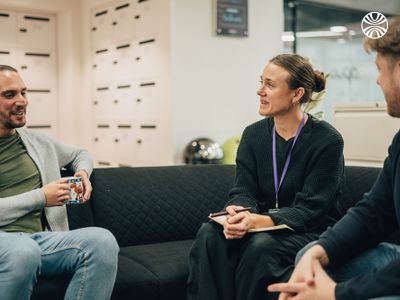 2 colleagues in casual conversation on a dark sofa in an office break area.