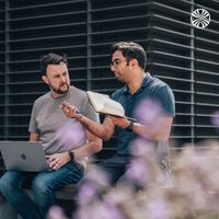 Two men with laptops sit on a bench outside, one holding a notebook. Soft focus flowers in the foreground.